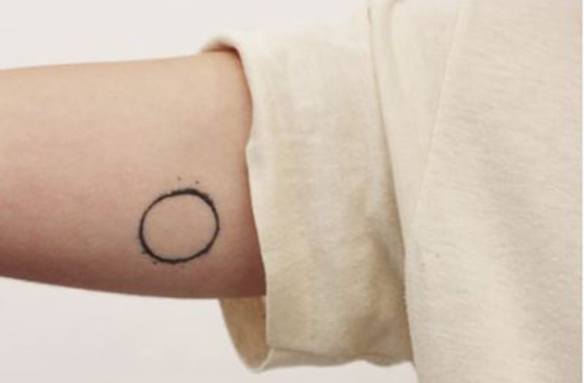 Um braço com tatuagem de círculo preto e pontos, próximo a uma camisa clara.