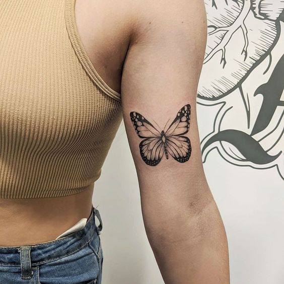 Pessoa com tatuagem de borboleta no bra&ccedil;o, vestindo uma blusa sem mangas bege.