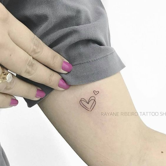 Pessoa com unhas pintadas de rosa, mostrando uma tatuagem de cora&ccedil;&otilde;es no bra&ccedil;o.