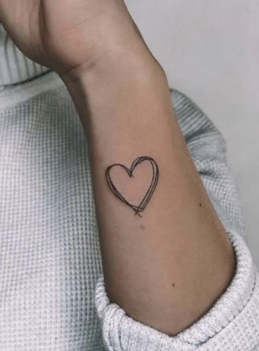 Bra&ccedil;o com tatuagem de cora&ccedil;&atilde;o de tra&ccedil;os finos no pulso, vestindo uma blusa cinza.