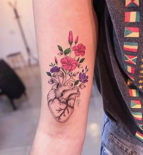 Tatuagem de coração anatômico com flores coloridas no antebraço de uma pessoa.
