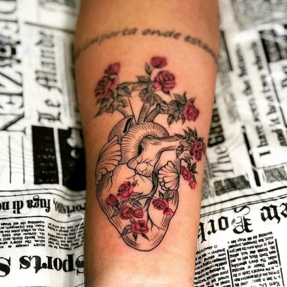 Uma tatuagem de coração anatômico com rosas vermelhas e folhas em um braço, sobre um fundo de jornal.