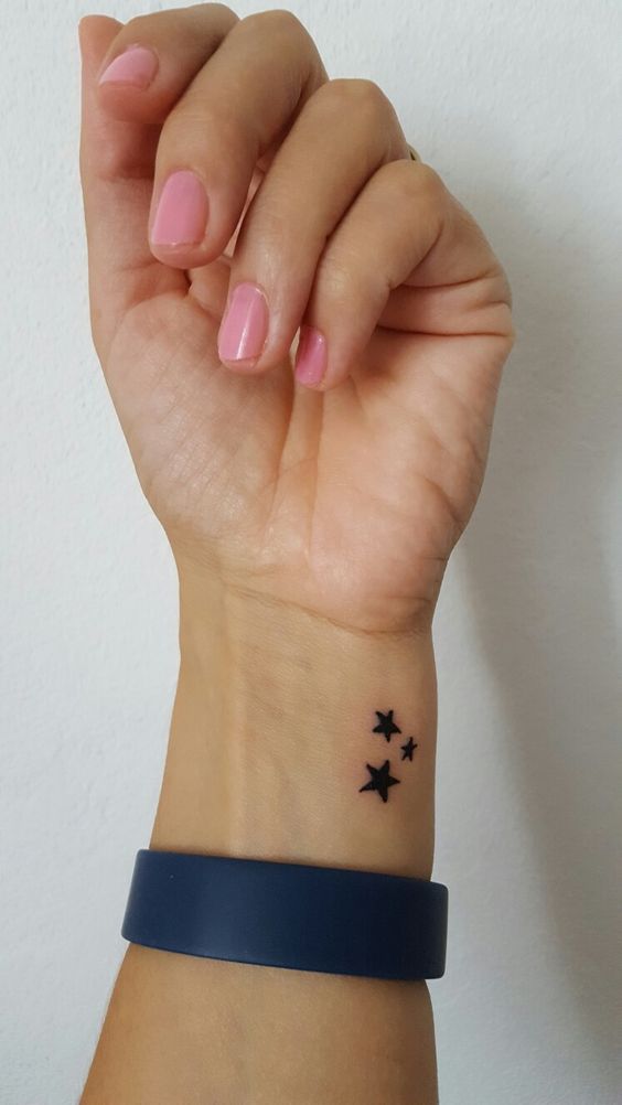 M&atilde;o com unhas pintadas de rosa e tatuagem de estrelas no pulso, usando uma pulseira azul escura.
