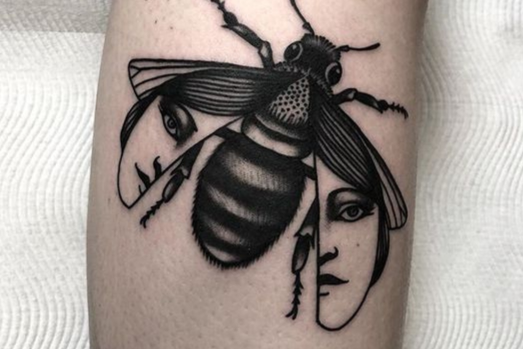 Tattoo de uma abelha com asas formando rostos, em estilo preto e cinza, sobre a pele.