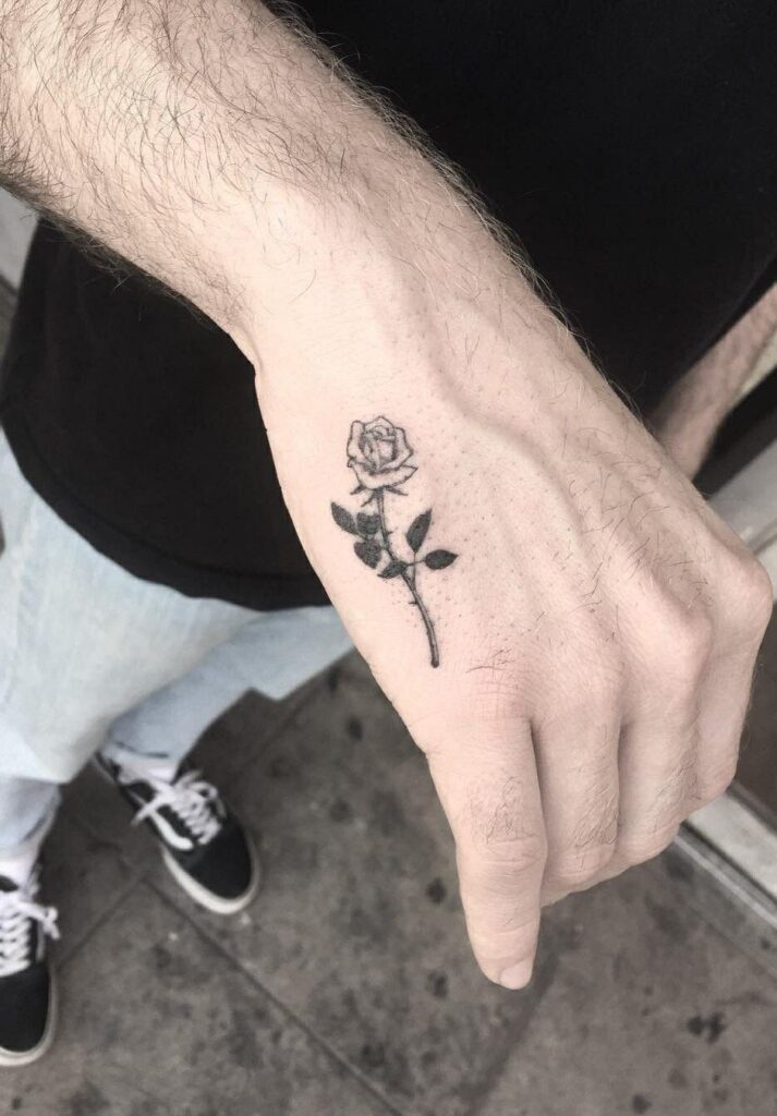 Mão com tatuagem de rosa preta no dorso, sobre fundo de calçada e pessoa usando calça clara e tênis preto.