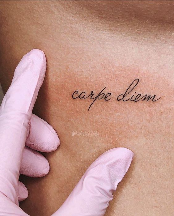 Uma tatuagem com a frase "carpe diem" é mostrada na pele, sendo tocada por mãos com luvas rosas.