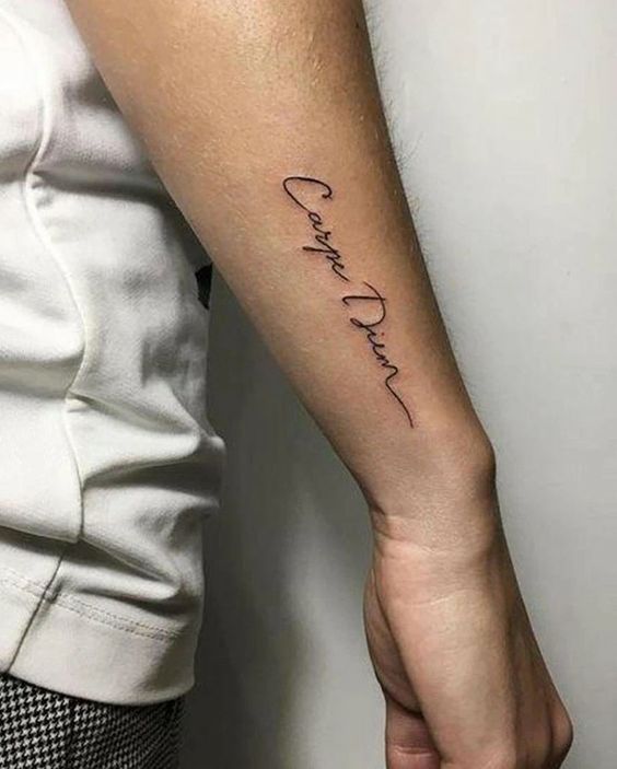 Braço com tatuagem "Carpe Diem" em letra cursiva, próximo ao cotovelo, usando camiseta branca.