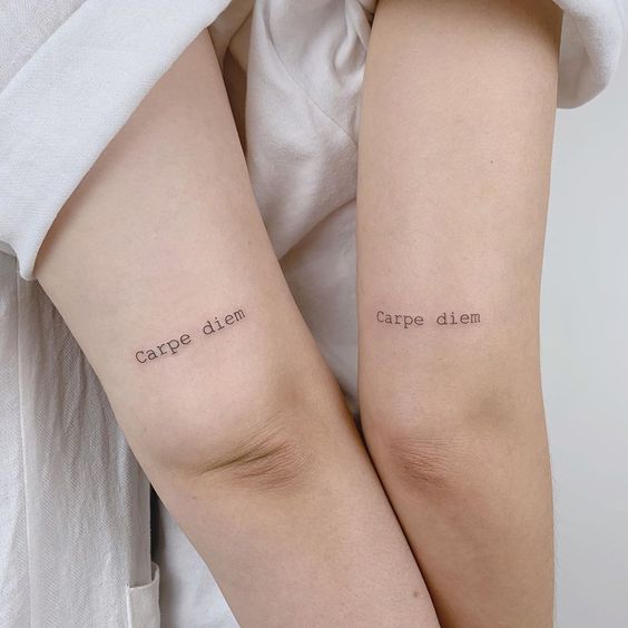 Dois braços com tatuagens idênticas "carpe diem" na parte interna, próximo ao cotovelo.