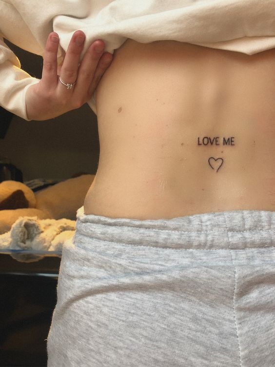 Pessoa com tatuagem "LOVE ME" e cora&ccedil;&atilde;o na lateral do abd&ocirc;men, vestindo cal&ccedil;a cinza e blusa clara.