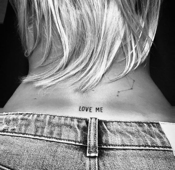 Cabelo loiro curto sobre uma tatuagem de constela&ccedil;&atilde;o e a frase "LOVE ME" na nuca de uma pessoa em jeans.