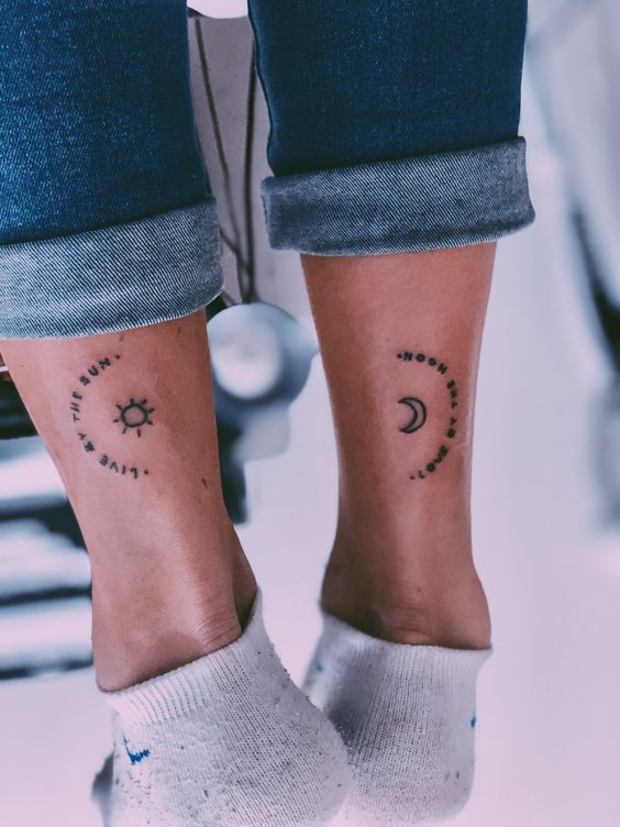 Calcanhares com tatuagens de sol e lua, rodeadas por frases, vis&iacute;veis acima de meias brancas e cal&ccedil;as dobradas.