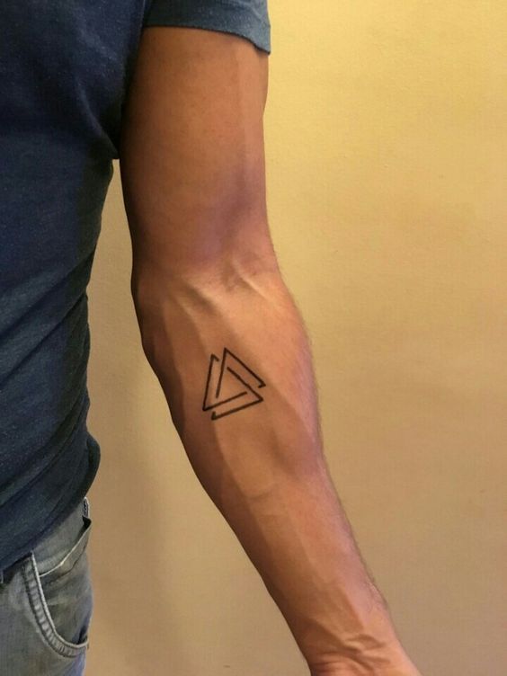 Bra&ccedil;o de uma pessoa com tatuagem geom&eacute;trica de tri&acirc;ngulos pretos no antebra&ccedil;o, vestindo camiseta azul.