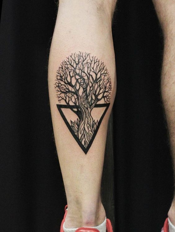 Tatuagem de &aacute;rvore estilizada dentro de um tri&acirc;ngulo invertido na perna de uma pessoa, com fundo escuro.