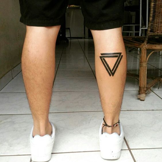 Pernas de uma pessoa com tatuagem geom&eacute;trica na panturrilha, usando t&ecirc;nis brancos e shorts pretos, em um piso claro.