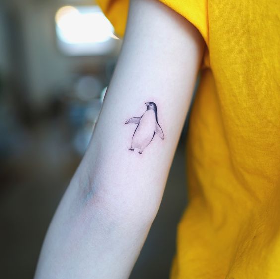 Pessoa com camiseta amarela exibe tatuagem de pinguim no braço.