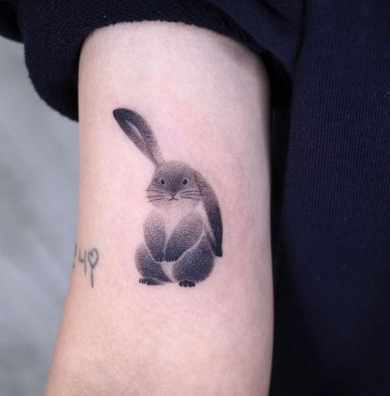 Tatuagem de coelho estilizado em tons de cinza no braço de uma pessoa usando blusa preta.