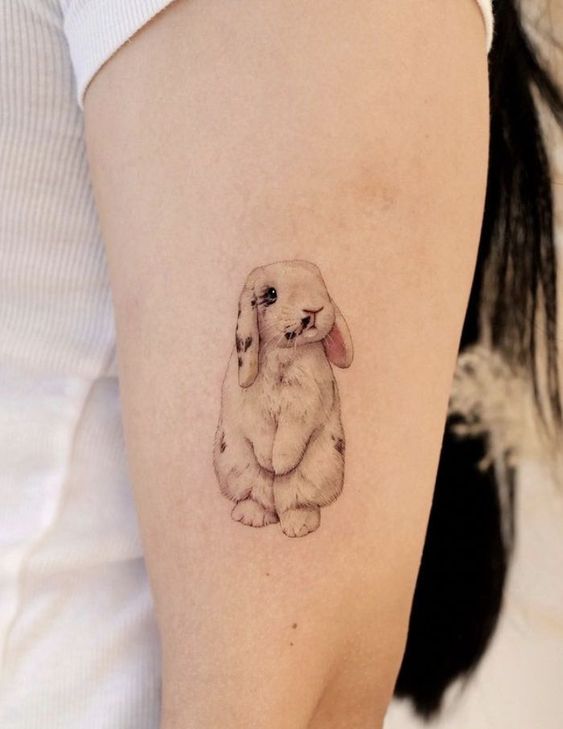 Tatuagem de coelho fofo no braço de uma pessoa com camiseta branca.