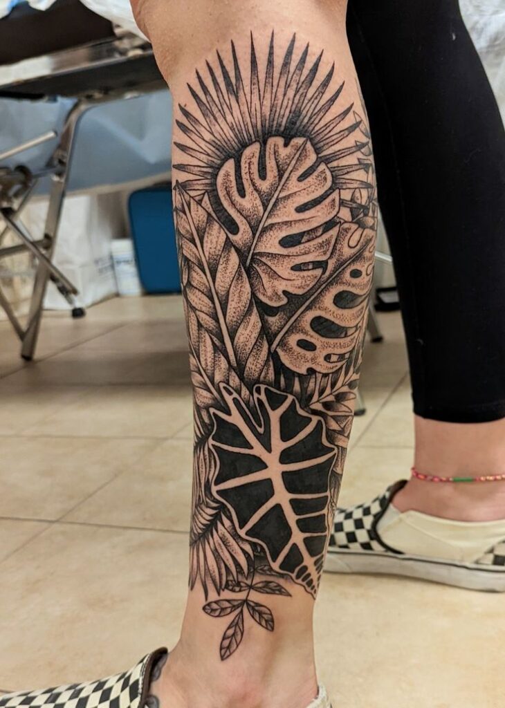 Tatuagem de folhas tropicais em preto e cinza na perna de uma pessoa, com detalhes de linhas e sombras.