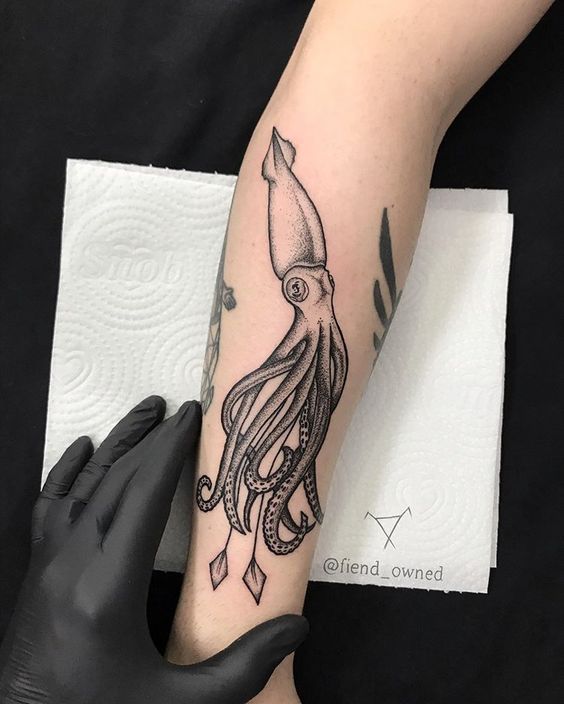 Bra&ccedil;o com tatuagem de lula detalhada em preto, mostrado sobre papel toalha, m&atilde;o com luva preta ao lado.