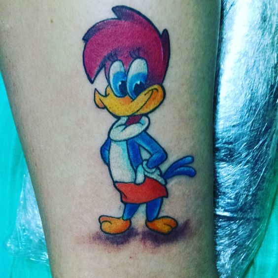 Tatuagem colorida de um p&aacute;ssaro animado com penas azuis e topete vermelho em um antebra&ccedil;o.
