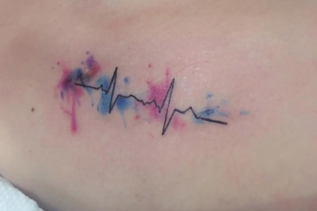 tatuagem-batimento-cardiaco-3-1024x683-1 Tatuagem de linha de eletrocardiograma em preto com manchas coloridas de tinta rosa e azul ao redor.