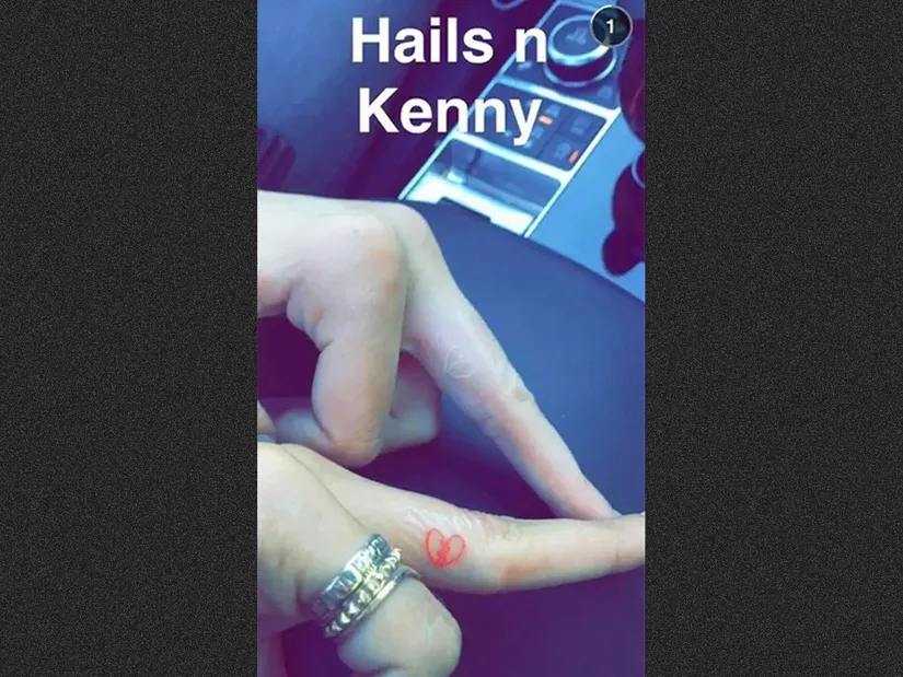 M&atilde;os com an&eacute;is mostram tatuagem de cora&ccedil;&atilde;o no dedo em ambiente interno, com parte de um painel de carro vis&iacute;vel ao fundo.