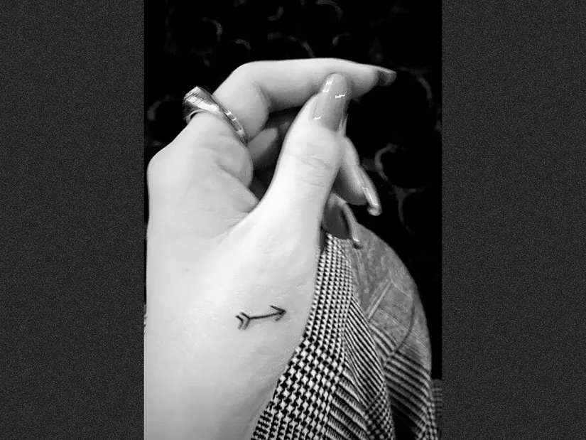 M&atilde;o segurando tecido, com tatuagem de flecha no dedo e anel prateado. Fundo escuro.