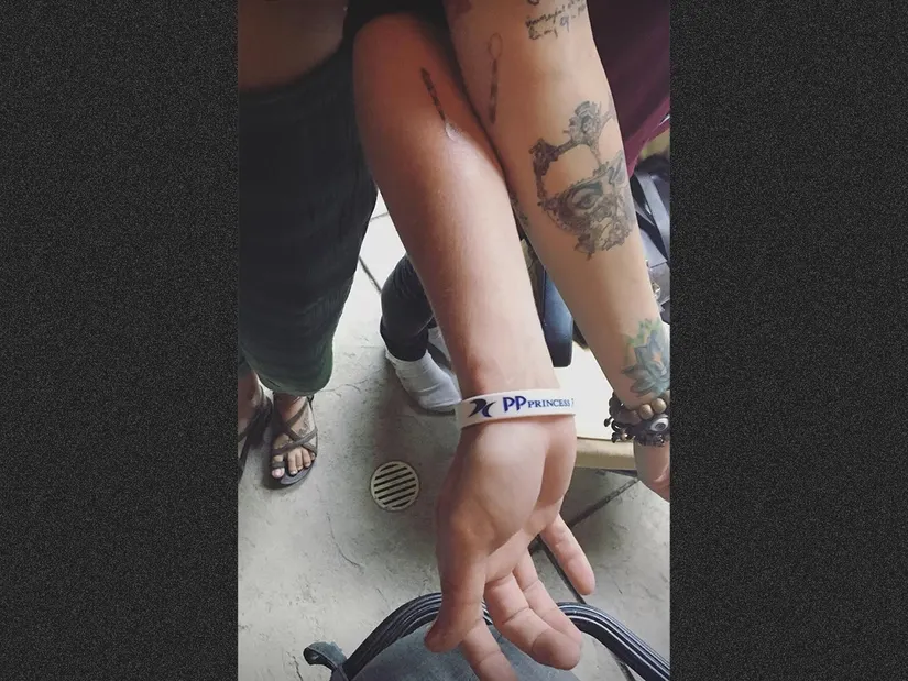 Dois bra&ccedil;os tatuados se tocam; um usa uma pulseira branca com texto azul.