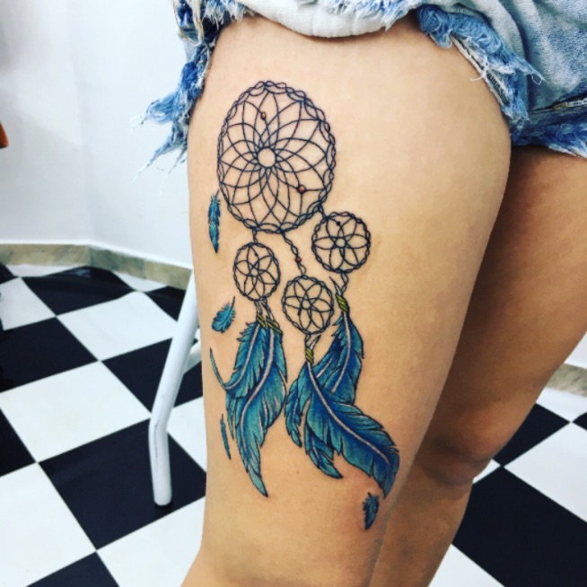 Tatuagem de filtro dos sonhos azul com penas na coxa de uma pessoa em um ambiente com piso preto e branco.