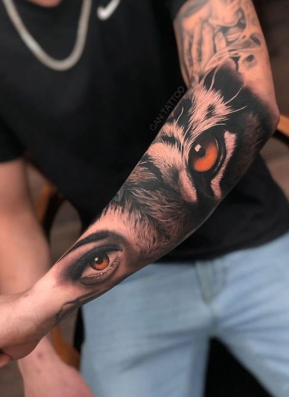 tatuagem-tigre-3 Um braço com tatuagens detalhadas de olhos humanos e de lobo, destacando-se em um fundo de roupa preta e jeans azul.