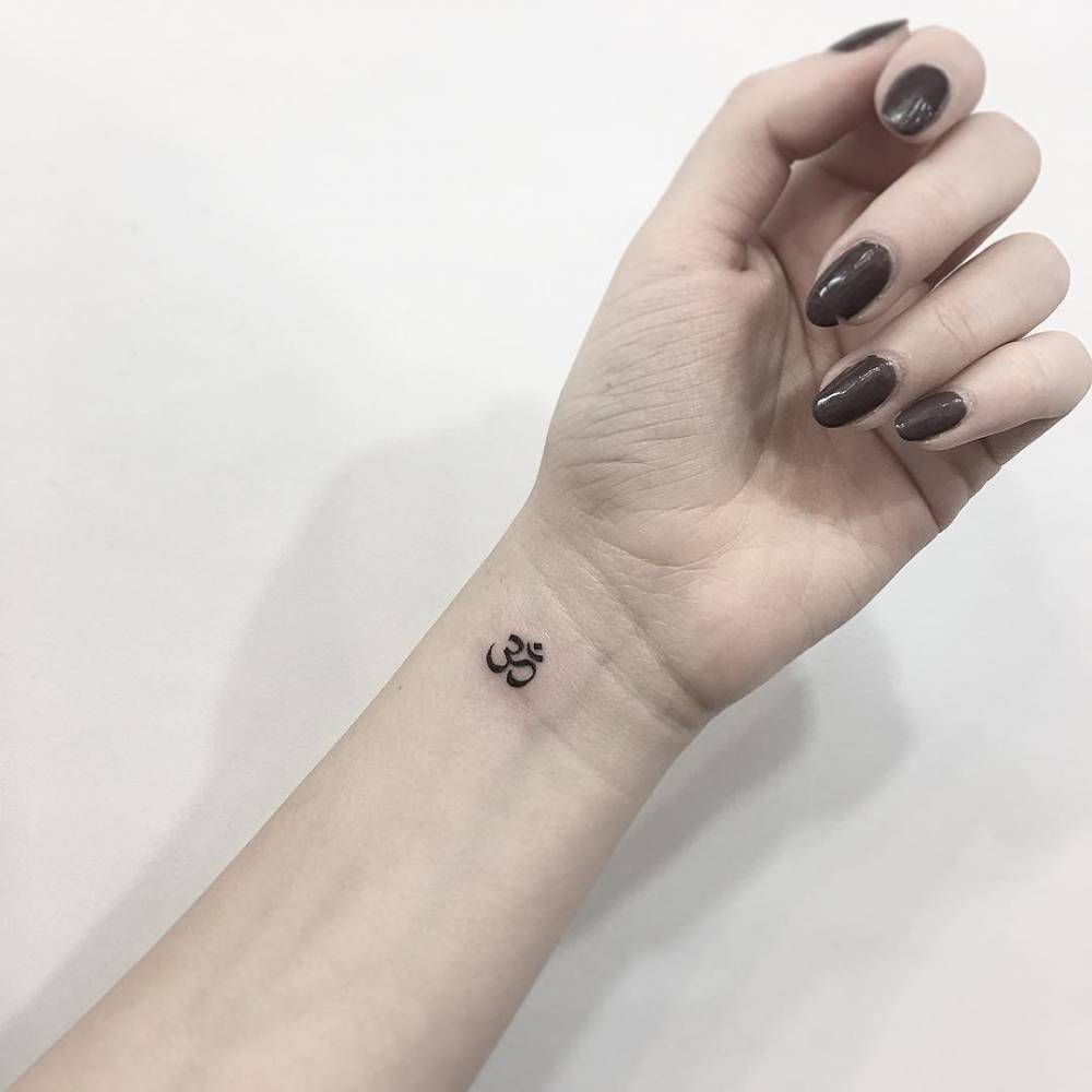 significado-tatuagem-om-2 Pulso com tatuagem de símbolo em preto, unha esmaltada em tom escuro. Fundo claro.
