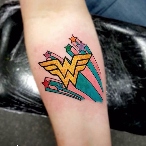 tatuagem-mulher-maravilha-2 Uma tatuagem colorida no braço com um emblema dourado e estrelas coloridas ao redor.