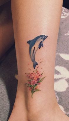significado-tatuagem-de-golfinho-2 Tatuagem de golfinho azul saltando sobre flores rosas no tornozelo de uma pessoa.