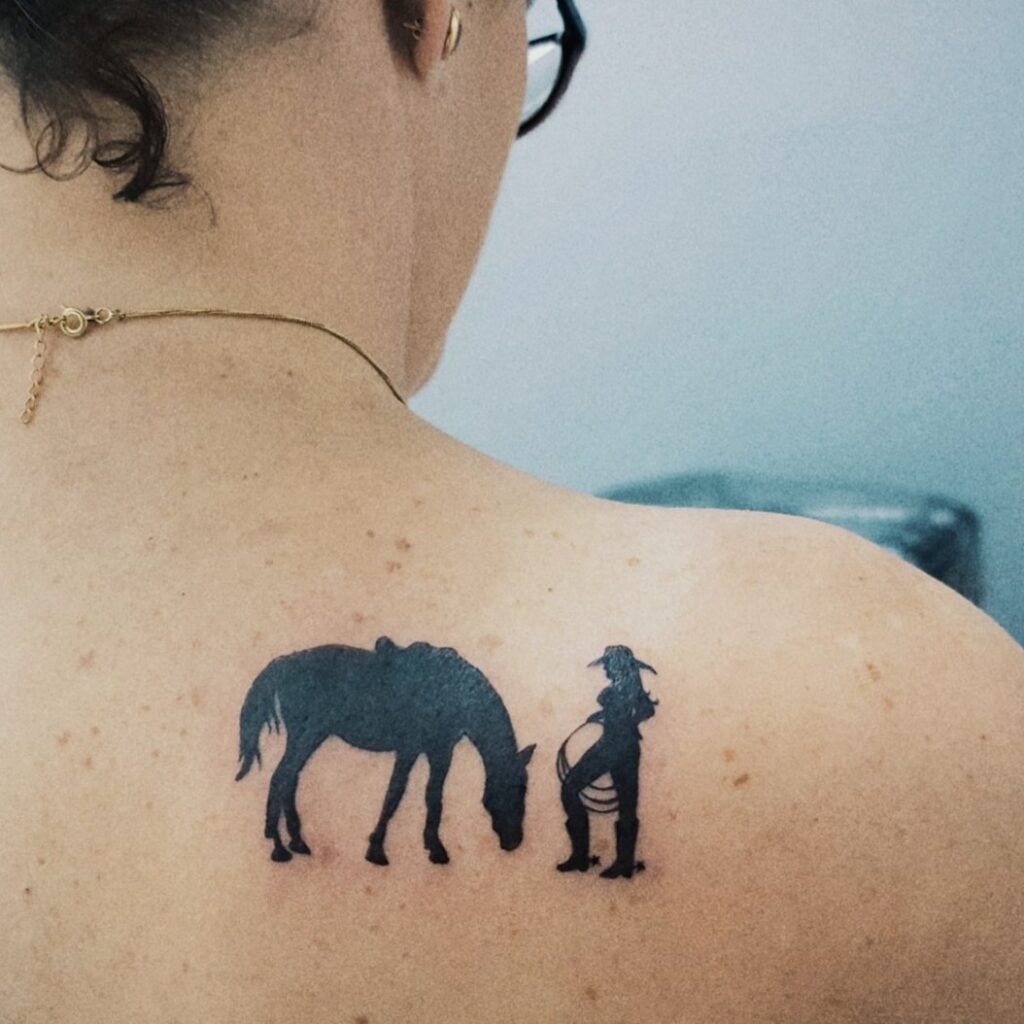 Uma tatuagem de silhueta de um cavalo e uma pessoa em um ombro.