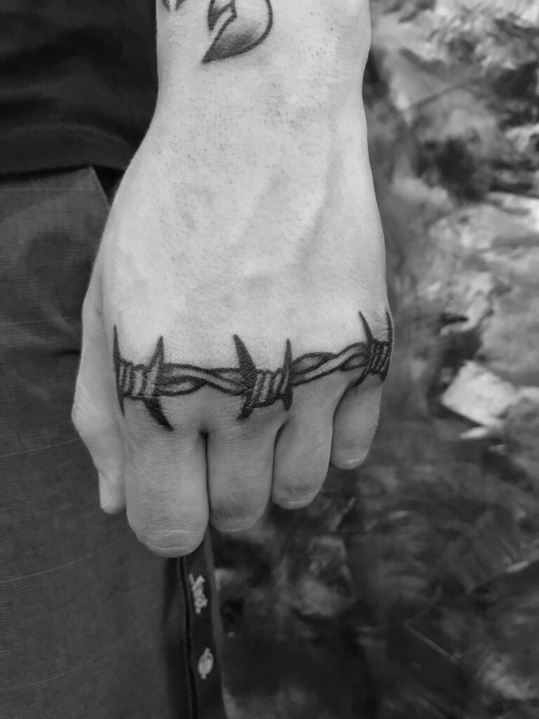 tatuagem-arame-farpado-2-768x1024-1 Uma mão com uma tatuagem de arame farpado no dorso, em uma imagem em preto e branco.