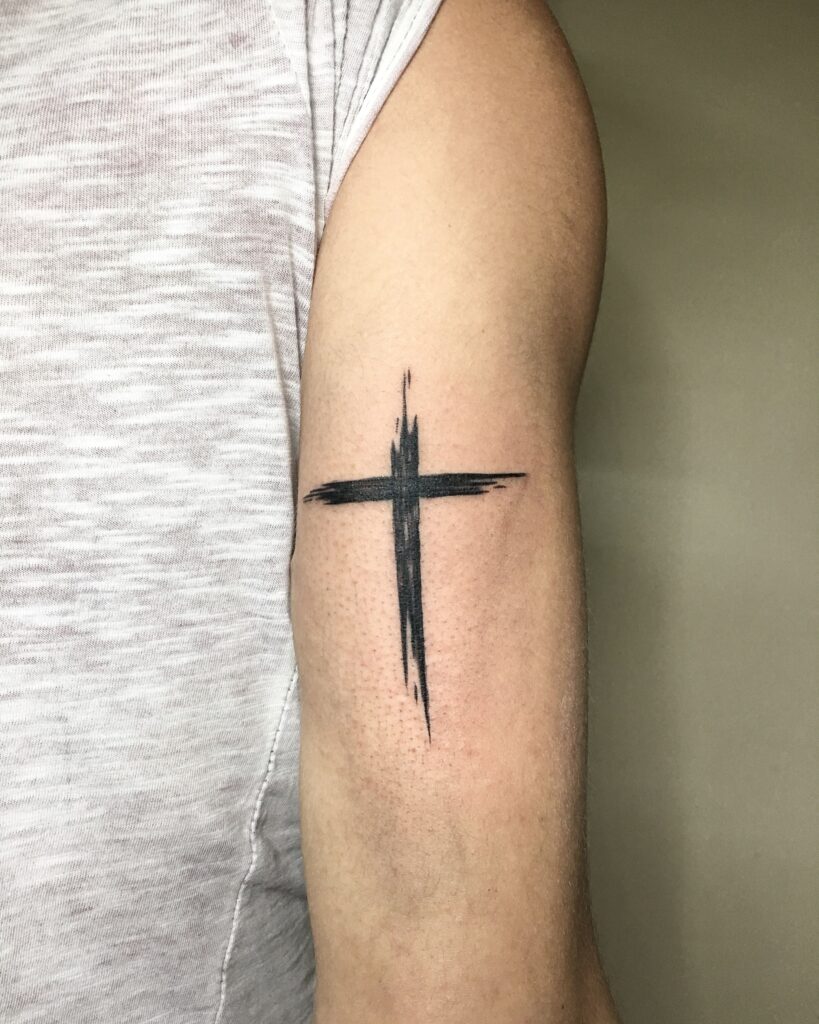 Bra&ccedil;o com tatuagem de cruz preta estilizada, vestindo camiseta cinza.