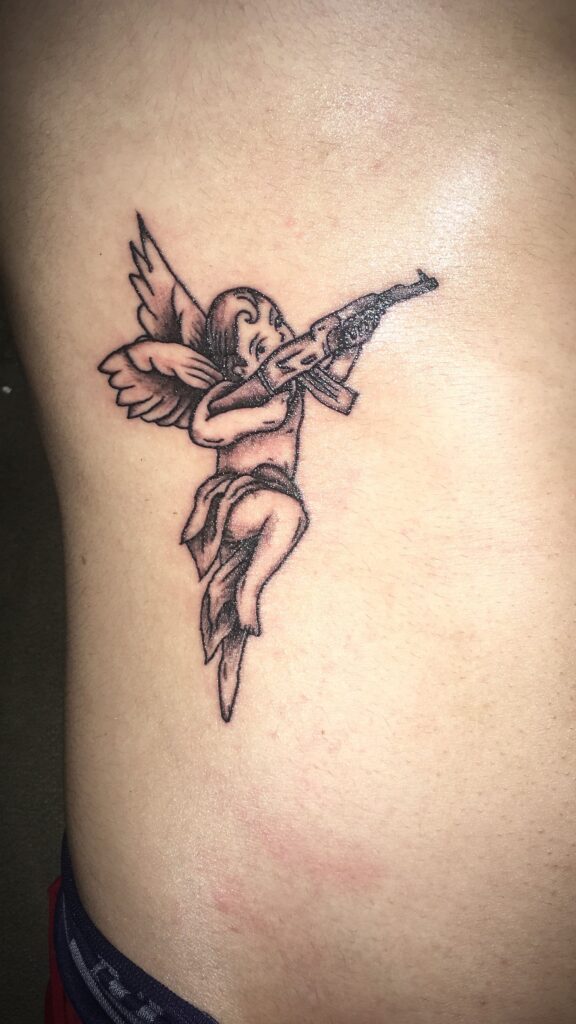 Tatuagem de anjo alado segurando um rifle, desenhado em pele clara.