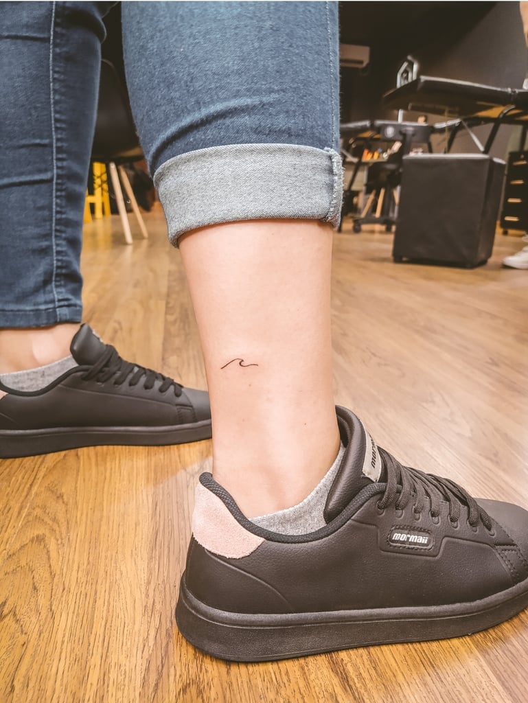 w769 Pessoa de calça jeans dobrada e tênis preto, exibindo uma pequena tatuagem no tornozelo em ambiente interno.