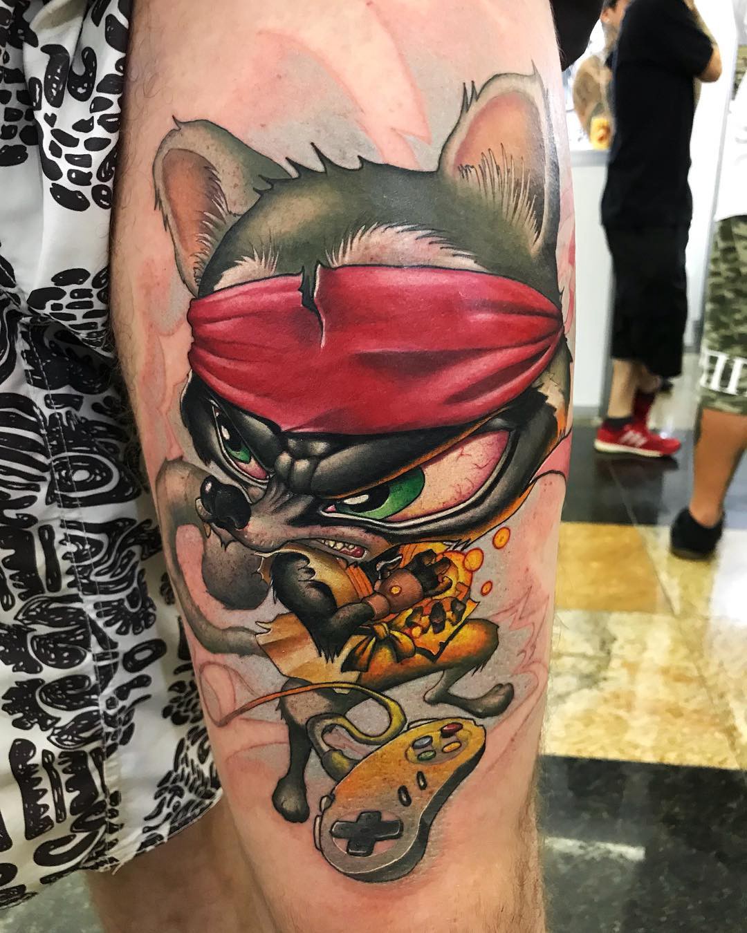 1gj2p91yr0gvgclcmbmrgow-1 Tatuagem de personagem animal com bandana vermelha, roupa amarela e controle de videogame no braço de uma pessoa.