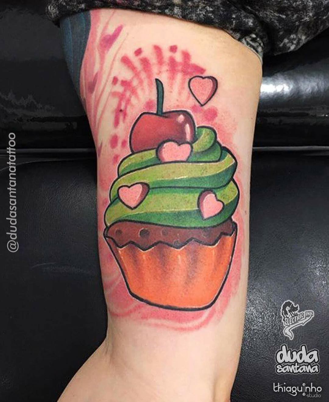 18nedlpdo3oz-spg_fytxka-1 Tatuagem de cupcake colorido com cobertura verde, cereja no topo e corações, no braço de uma pessoa.
