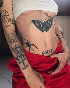 67e1fd8988e12a8ca39cd5c04d5e554a Pessoa com várias tatuagens no corpo, incluindo borboleta e aranha, vestindo top branco e calça vermelha.