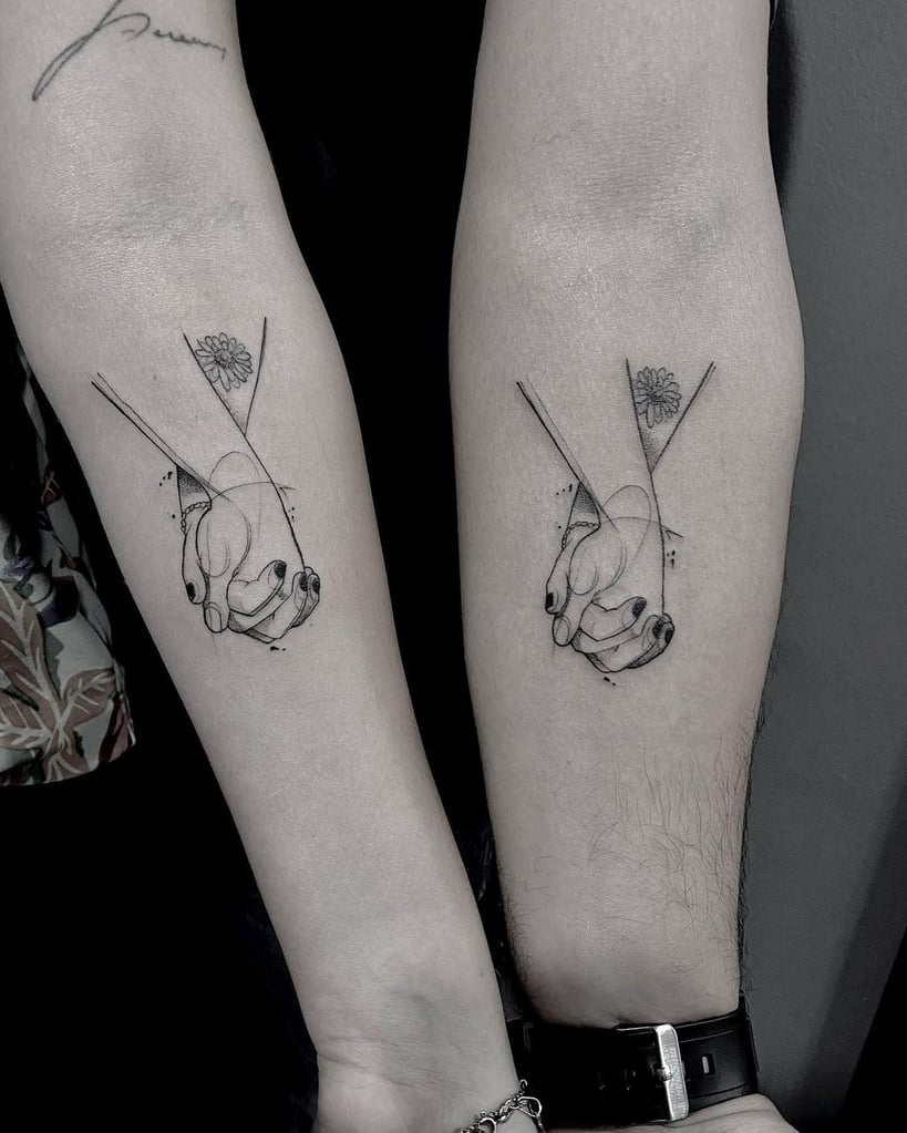 w819-179 Duas tatuagens nos antebraços exibem mãos entrelaçadas e uma flor, com linhas geométricas em estilo minimalista.