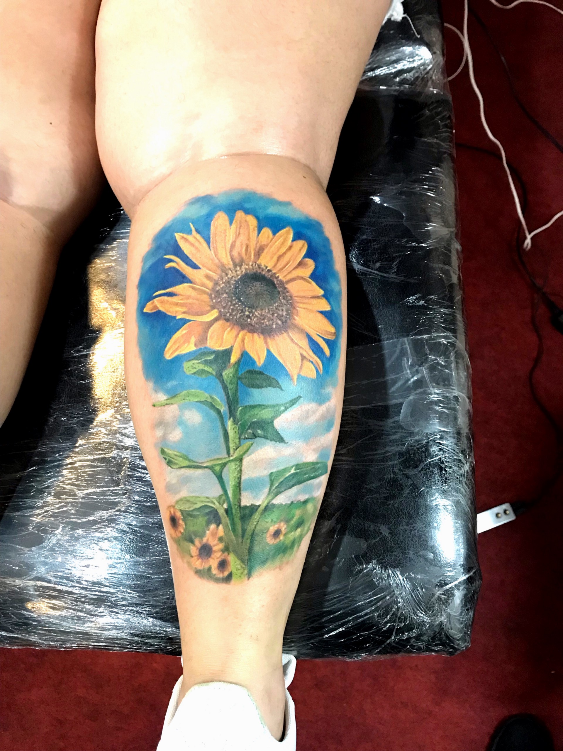 1s52wjg92ye9-miyq5muqka Perna com tatuagem colorida de girassol em um fundo azul.