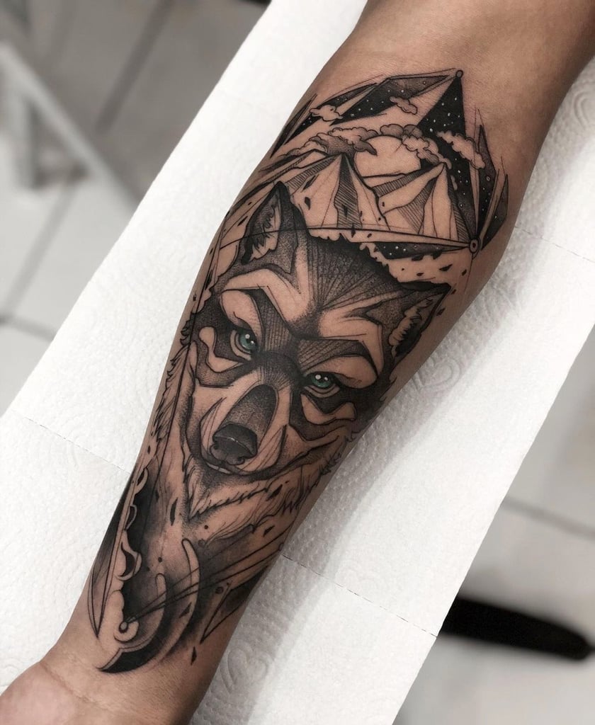 Tatuagem intricada de lobo com montanhas e céu estrelado no braço de uma pessoa, arte em preto e cinza.
