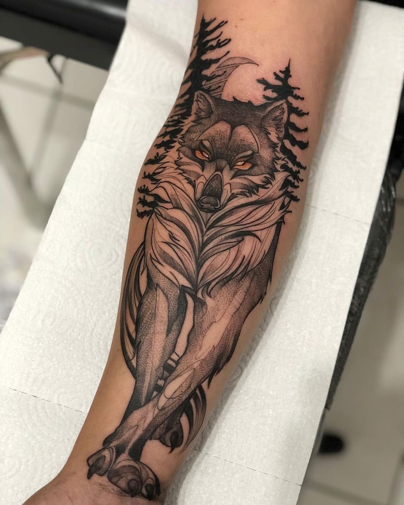 Tatuagem de lobo estilizado com árvores e lua ao fundo, localizada no antebraço de uma pessoa.