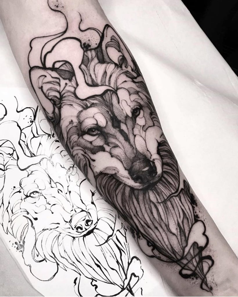 Braço tatuado com desenho de lobo detalhado em preto, acompanhado de esboço do mesmo lobo ao lado.