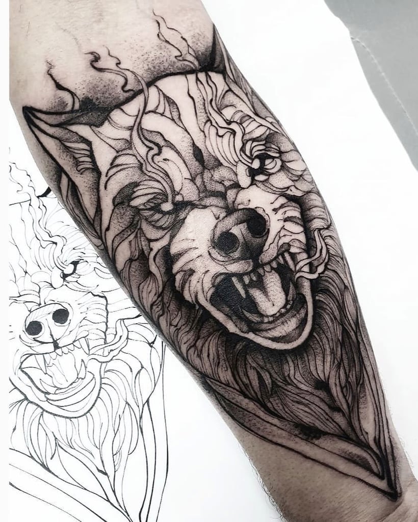 Tatuagem no braço de um lobo detalhado em preto, acompanhado de um esboço similar no papel ao lado.