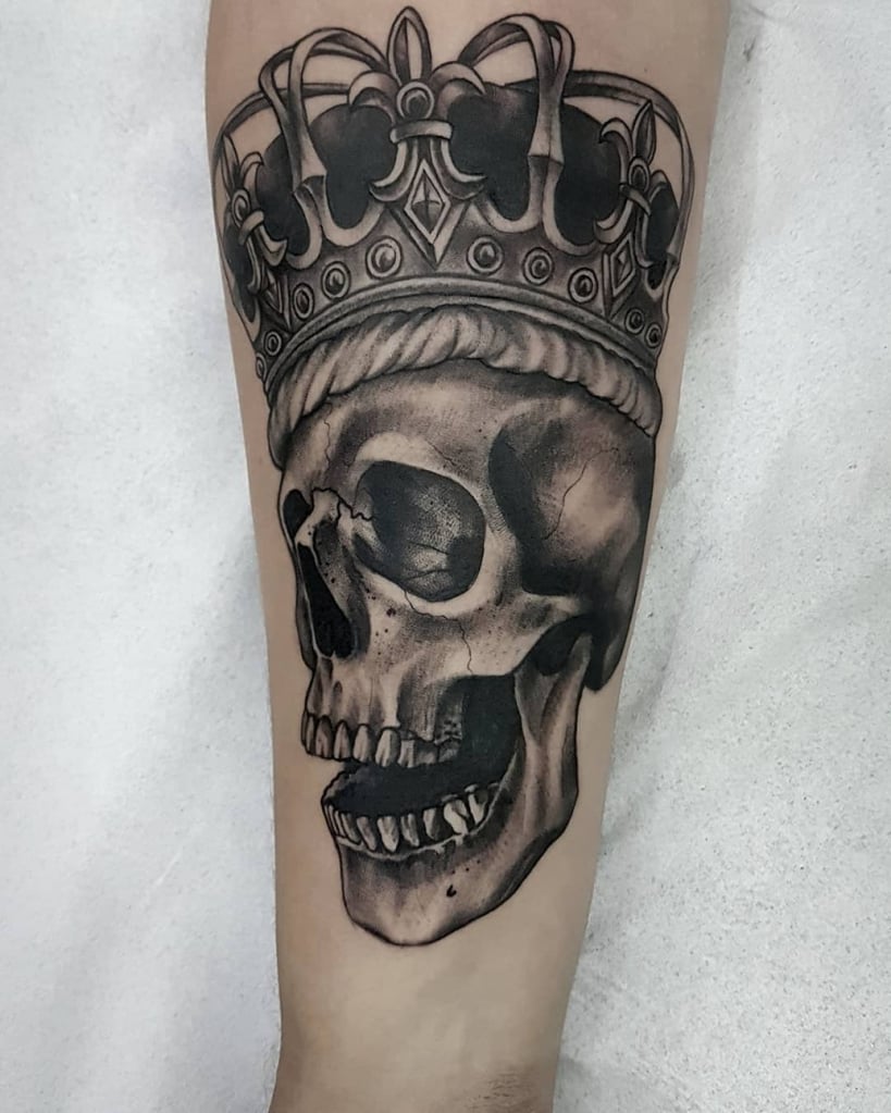 w819-324 Tatuagem de caveira com coroa detalhada, em tons de preto e cinza, no braço de uma pessoa.