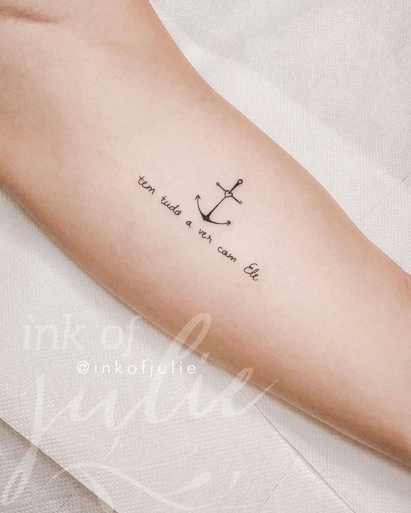 w819-328 Braço com tatuagem de âncora e frase "tem tudo a ver com Ele" em letras cursivas.
