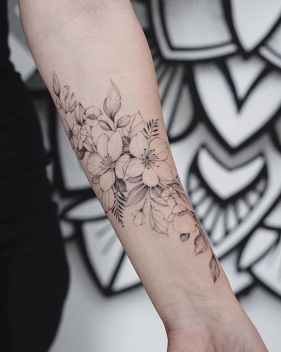 Tatuagem no antebraço com desenho delicado de flores e folhas em preto e cinza, destacando arte floral detalhada.